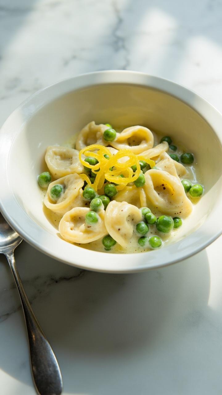 white shallow bowl with pale cream sauce coating plump tortellini, scattered bright green peas, thin lemon zest strips on top, small spoon to the side, marble countertop white shallow bowl with pale cream sauce coating plump tortellini, scattered bright green peas, thin lemon zest strips on top, small spoon to the side, marble countertop