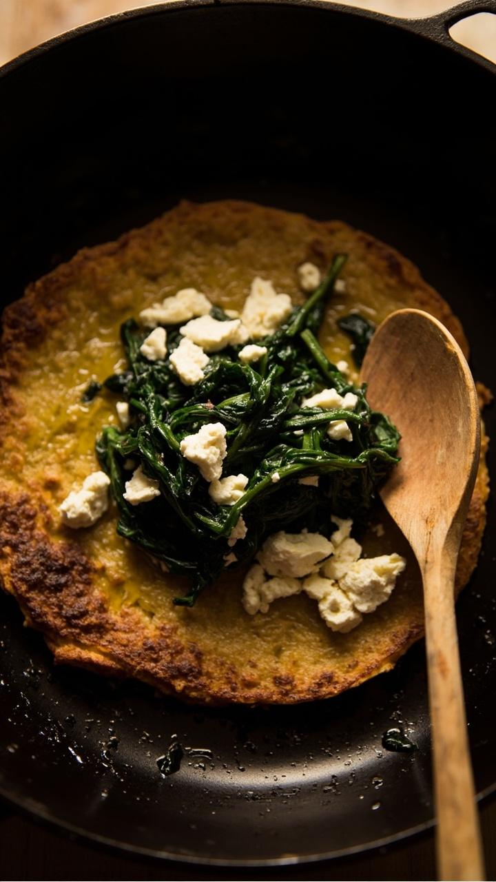 a round golden socca base topped with wilted spinach, scattered feta crumbles, a light drizzle of olive oil, served on a cast iron pan with a wooden spoon beside it a round golden socca base topped with wilted spinach, scattered feta crumbles, a light drizzle of olive oil, served on a cast iron pan with a wooden spoon beside it