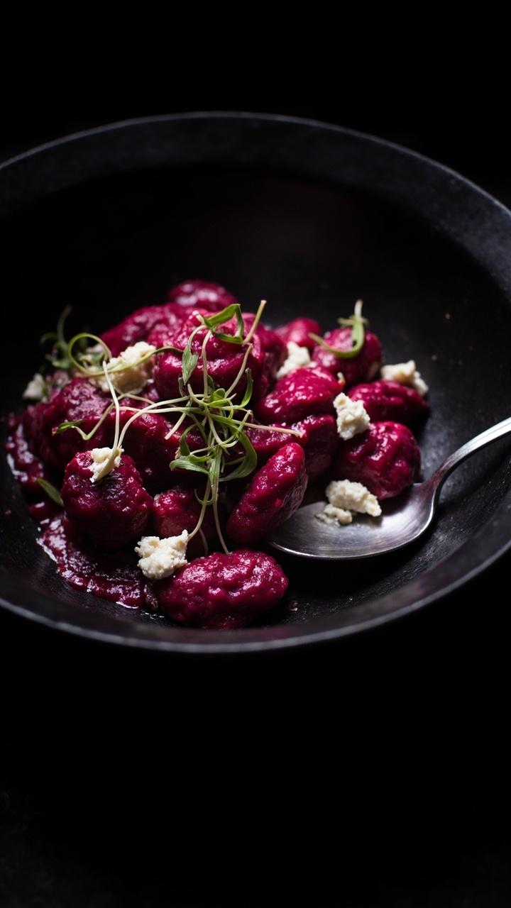 A shallow black bowl with bright magenta beet gnocchi lightly sauced and dotted with crumbled white goat cheese. Microgreens scattered on top for contrast. A small spoon at the side of the bowl. A shallow black bowl with bright magenta beet gnocchi lightly sauced and dotted with crumbled white goat cheese. Microgreens scattered on top for contrast. A small spoon at the side of the bowl.