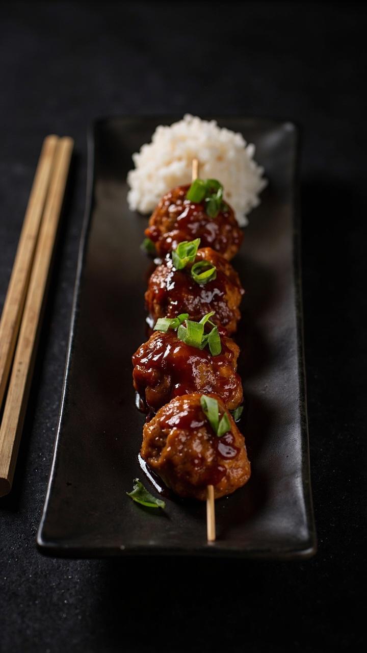 A dark rectangle plate with five glazed pork meatballs on skewers, thinly sliced scallions scattered across, a small mound of steamed rice at one end, a pair of bamboo picks beside the plate A dark rectangle plate with five glazed pork meatballs on skewers, thinly sliced scallions scattered across, a small mound of steamed rice at one end, a pair of bamboo picks beside the plate
