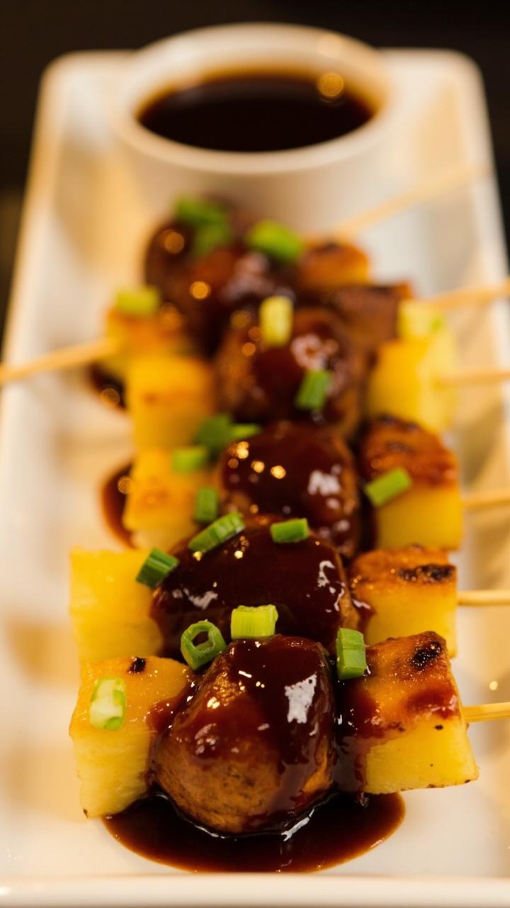 Glazed golden chicken meatballs with small pineapple chunks skewered on bamboo picks, glossy dark teriyaki glaze, sliced green onion sprinkled over, served on a long white platter with a small bowl of extra glaze
