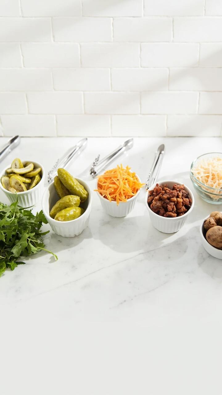 A countertop lined with five small bowls each with a different topping sliced dill pickles shredded cheddar crispy bacon arugula and grilled mushrooms a clean white tile backsplash and small tongs for each bowl