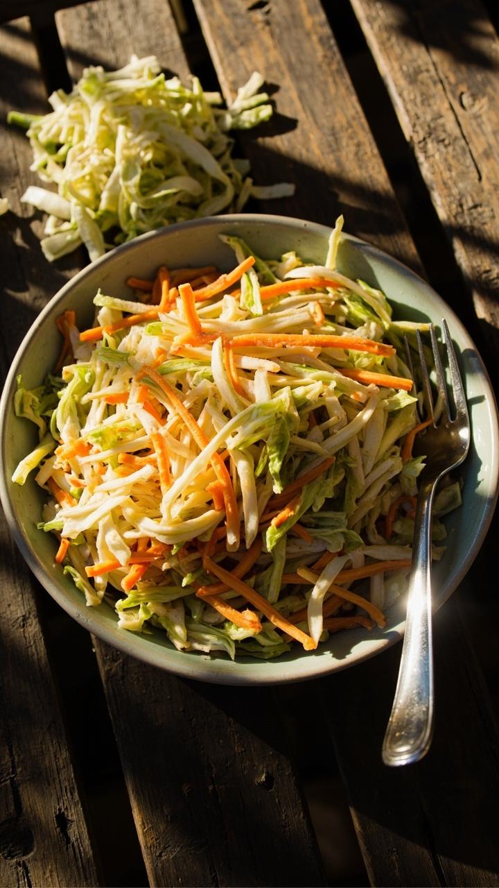 A shallow bowl of shredded cabbage and carrot slaw with a glossy dressing, a small pile of extra shredded cabbage on the side, a fork resting on the bowl rim, sunlight on a picnic table