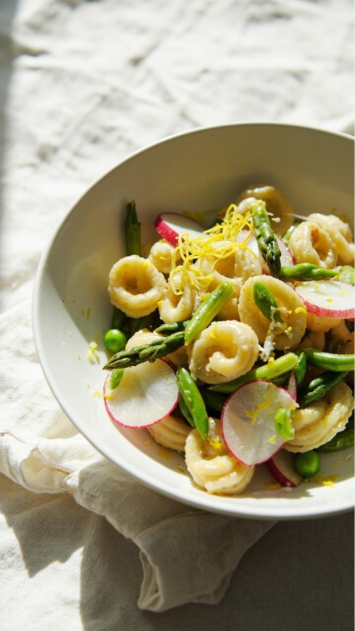 White shallow bowl with tortellini tossed with blanched asparagus tips, peas, sliced radishes, and a light olive oil dressing garnished with lemon zest on a pale cloth napkin